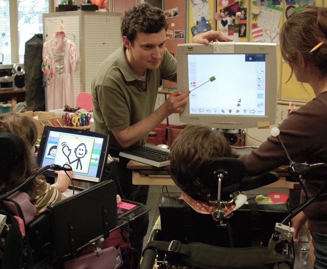 Anthony Hornof points to a computer screen with a small stick. Two girls in wheelchairs are facing the computer display.