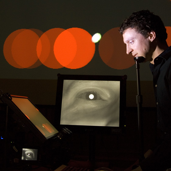 Anthony Hornof's face illuminated in a dark room, surrounded by computer displays showing big red circles bouncing around and images of his eyes. 